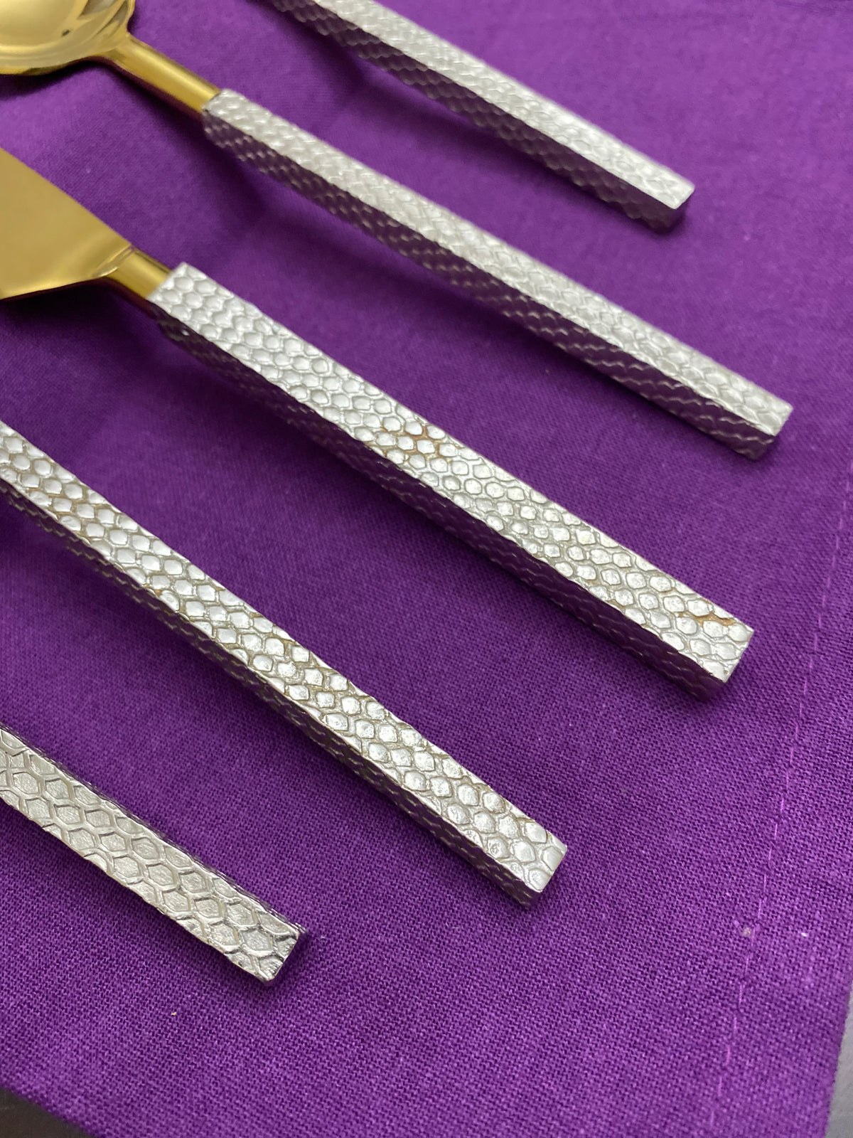 Close-up of five designer flatware pieces with hammered silver handles and gold-toned heads arranged on a purple fabric surface.