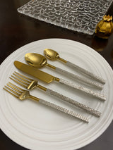 Golden stainless steel flatware set on a white round plate, featuring textured silver handles with a scaled pattern including a knife, two spoons, and two forks.