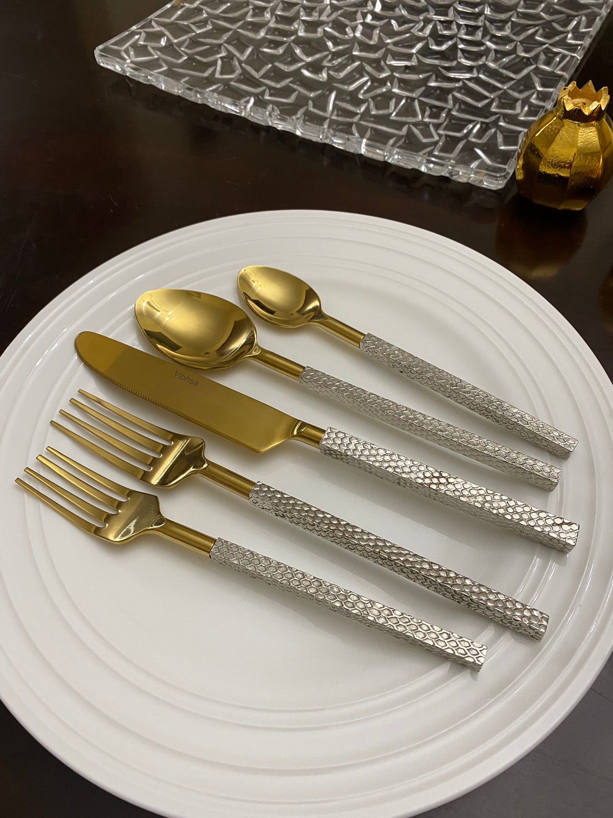 Golden stainless steel flatware set on a white round plate, featuring textured silver handles with a scaled pattern including a knife, two spoons, and two forks.