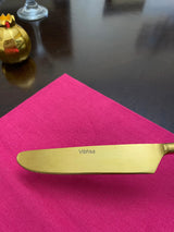 Golden stainless steel butter knife with serrated edge and 'Vibhsa' engraved on blade, resting on vibrant pink cloth against dark reflective surface.