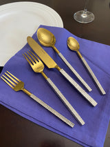 Flatware set with gold-colored heads and textured silver handles arranged on a purple napkin beside a white plate and wine glass base on a dark surface.