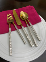 Gold-toned flatware with textured silver handles arranged on a white plate with a folded deep pink napkin beneath the knife and fork, shown from above.