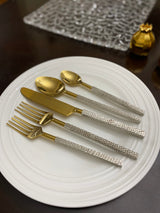 Golden stainless steel flatware set including two forks, a knife, and two spoons with textured silver handles arranged on a white round plate.