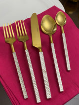 Five-piece gold flatware set with textured silver handles arranged on a folded deep pink napkin on a dark surface, including two forks, a knife, and two spoons.