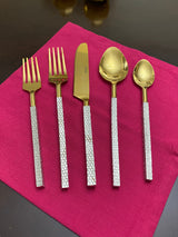 Golden stainless steel flatware with textured silver handles, arranged on a magenta cloth, including fork, knife, spoon, and teaspoon.