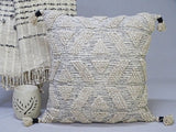 Beige woven throw pillow with geometric diamond patterns and textured cream tassels on each corner, placed beside a fringed woven blanket and a carved ceramic candle holder.