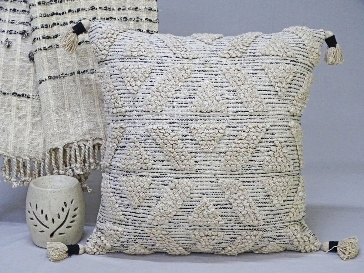 Beige woven throw pillow with geometric diamond patterns and textured cream tassels on each corner, placed beside a fringed woven blanket and a carved ceramic candle holder.