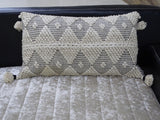 Rectangular cream-colored decorative throw pillow with black diamond and dot woven pattern, featuring textured knots and tassels on four corners, resting on a light textured surface.