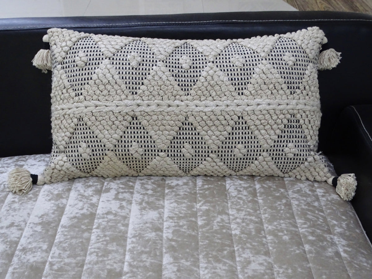 Rectangular cream-colored decorative throw pillow with black diamond and dot woven pattern, featuring textured knots and tassels on four corners, resting on a light textured surface.