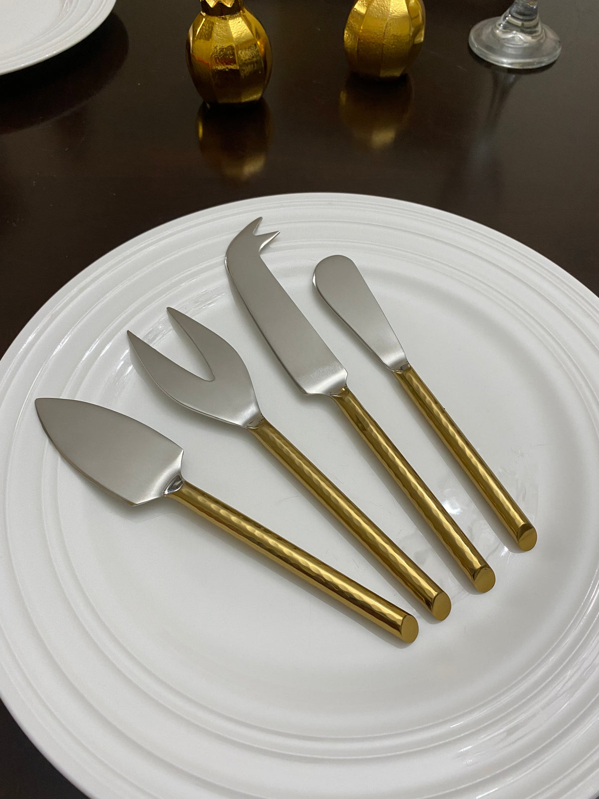 Set of four cheese knives with gold-colored handles and silver stainless steel blades arranged on a stacked white ceramic plate with concentric ridges.