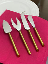 Set of four cheese knives with hammered gold handles and stainless steel blades, arranged on a bright pink cloth next to a white plate on a dark surface.
