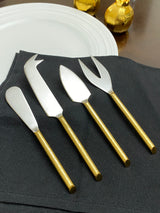 Set of four cheese knives with matte silver blades and shiny gold handles, arranged on a black cloth beside a white plate and golden foil-wrapped chocolates.