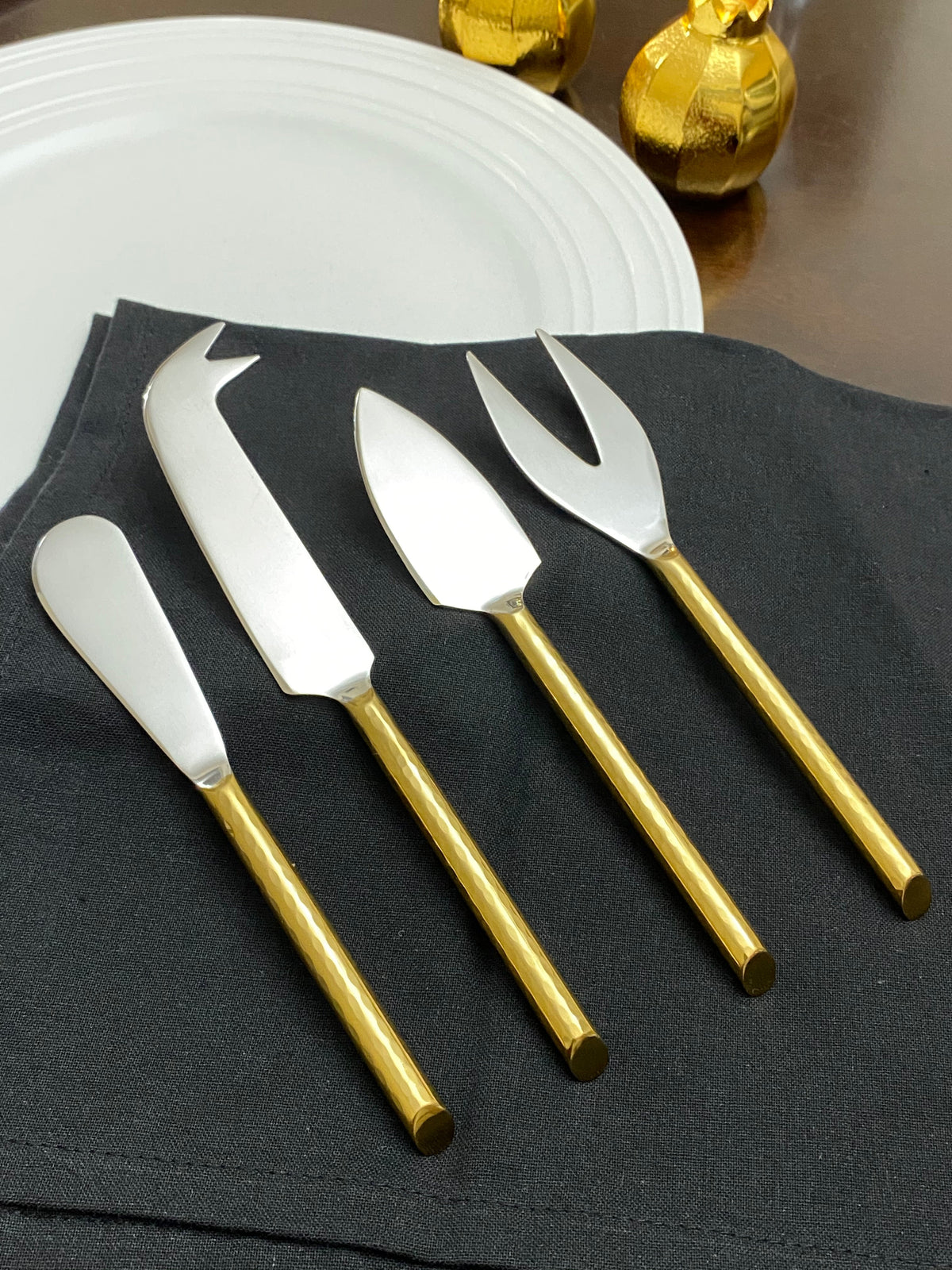 Set of four cheese knives with matte silver blades and shiny gold handles, arranged on a black cloth beside a white plate and golden foil-wrapped chocolates.