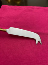 Cheese knife with a flat, slightly curved stainless steel blade ending in two pointed prongs and a gold-colored handle, placed on a bright pink fabric surface.