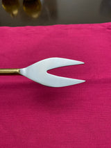 Stainless steel two-pronged cheese fork with a slender golden handle resting on a bright pink cloth surface.