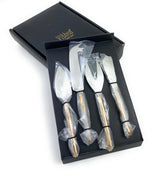 Set of four stainless steel cheese knives with gold-colored handles, each individually wrapped in plastic, displayed inside a black boxed packaging.