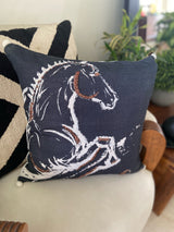 Square black decorative pillow with an embroidered white and brown horse outline design, placed on a chair with a black and beige geometric patterned pillow behind it.