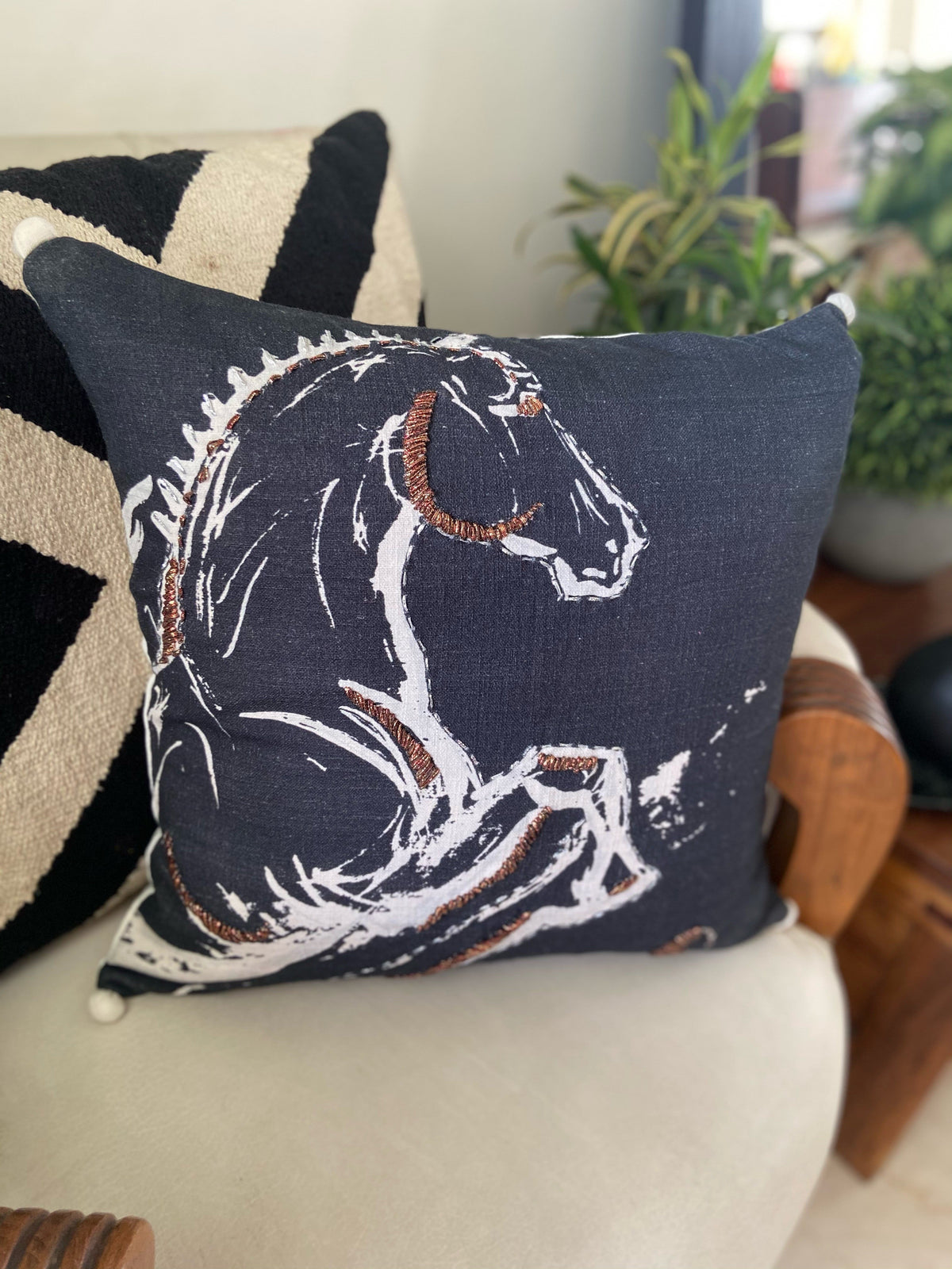 Square black decorative pillow with an embroidered white and brown horse outline design, placed on a chair with a black and beige geometric patterned pillow behind it.