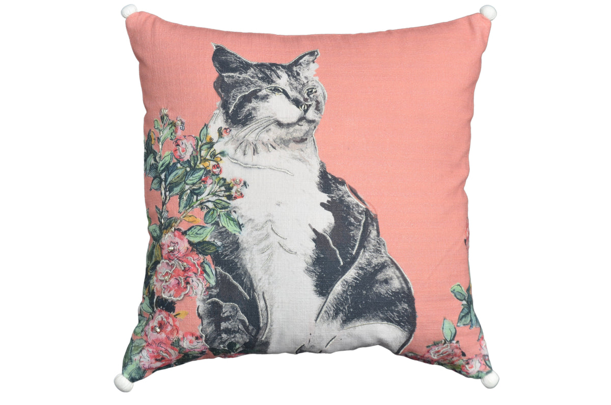 Square pink decorative pillow with a black and white cat illustration surrounded by pink flowers and green leaves, featuring white pom-poms at each corner.