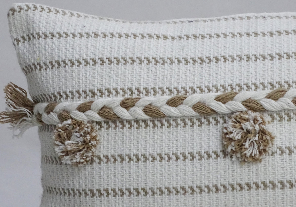 White throw pillow with light brown horizontal stripes and braided design topped with matching pom-poms on a neutral background.
