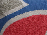 Close-up of a textured embroidery throw pillow featuring curved shapes in red, blue, beige, and gray yarns with a looped, tufted surface.