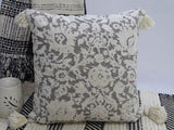 Square decorative throw pillow with tassels at corners, textured front fabric, placed on gray textured sofa.