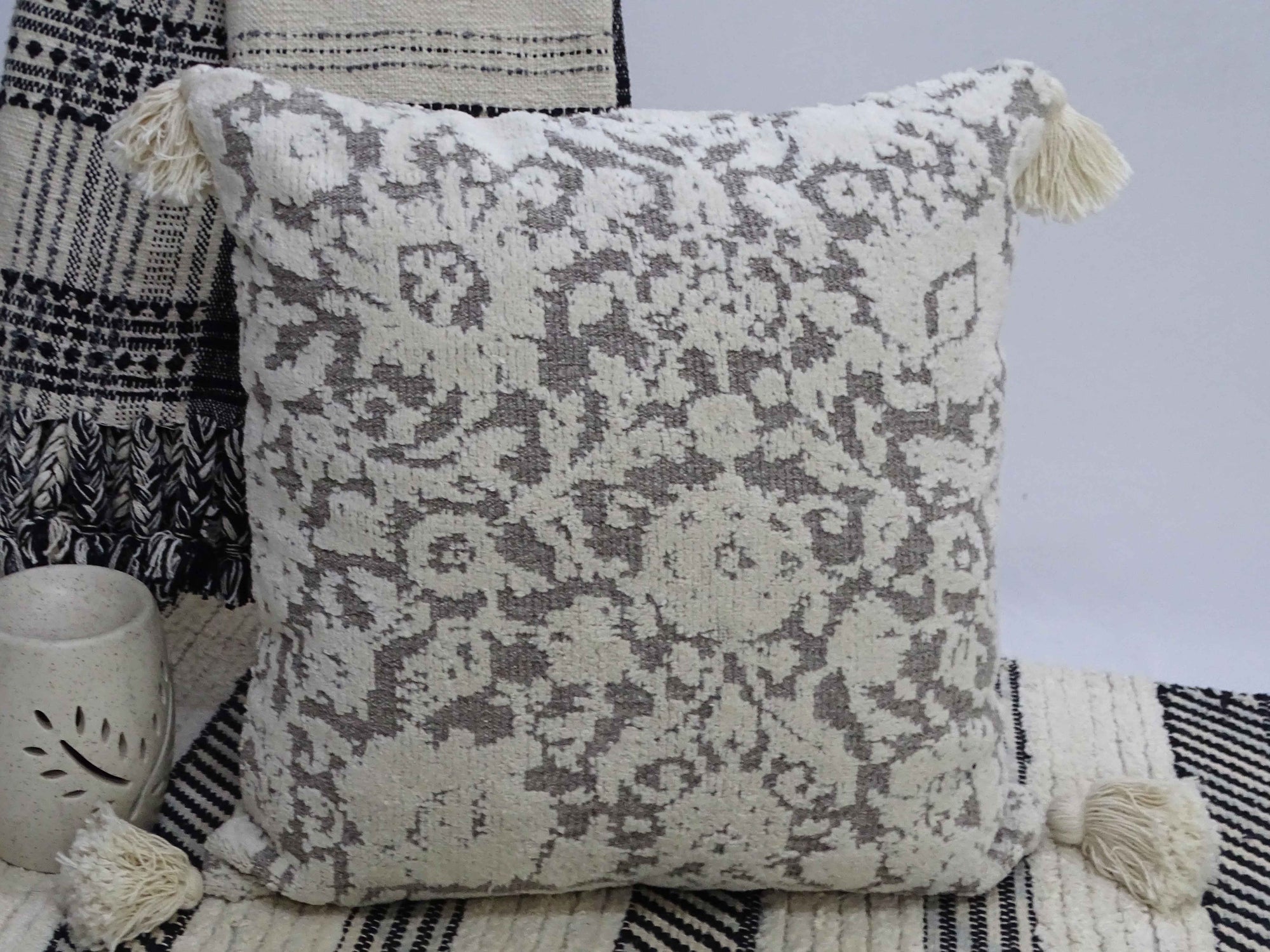 Square decorative throw pillow with tassels at corners, textured front fabric, placed on gray textured sofa.