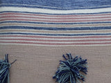 Close-up of a textured woven throw pillow with horizontal stripes in blue, white, and muted pink, featuring navy and light blue yarn pom-poms as accents.