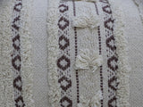 Close-up of handwoven cream-colored throw pillow with vertical brown geometric lines, textured fringe stripes, and three small horizontal fringe clusters.