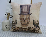 Beige throw pillow embroidered with a skull wearing a tall top hat, flanked by two black birds and a scroll banner reading 'Trick or Treat' at the bottom.
