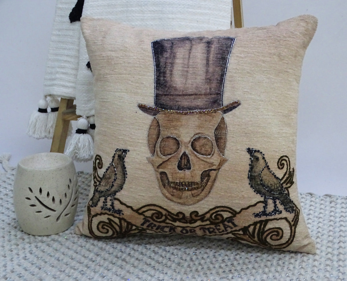Beige throw pillow embroidered with a skull wearing a tall top hat, flanked by two black birds and a scroll banner reading 'Trick or Treat' at the bottom.