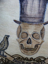 Close-up of a Halloween-themed embroidered throw pillow showing a brown skull wearing a dark top hat next to a black bird, with decorative brown scrollwork below.