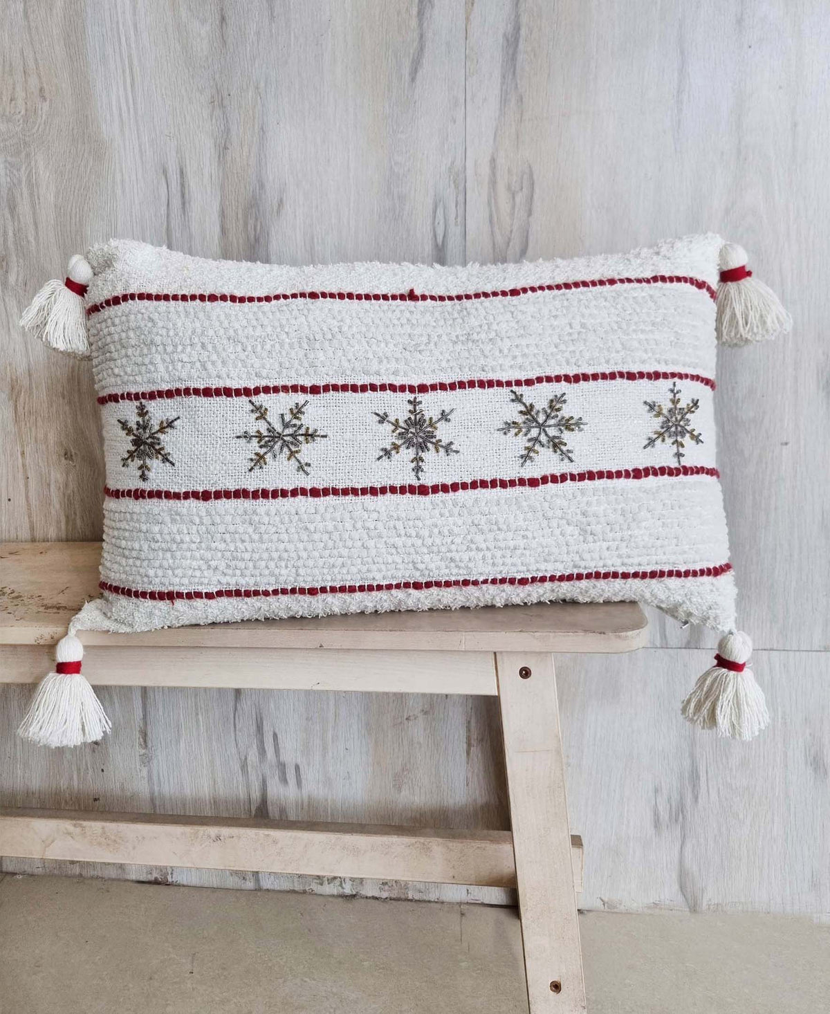 White rectangular pillow with red horizontal stripes and embroidered gray snowflakes, adorned with white tassels at each corner, resting on a light wooden bench.