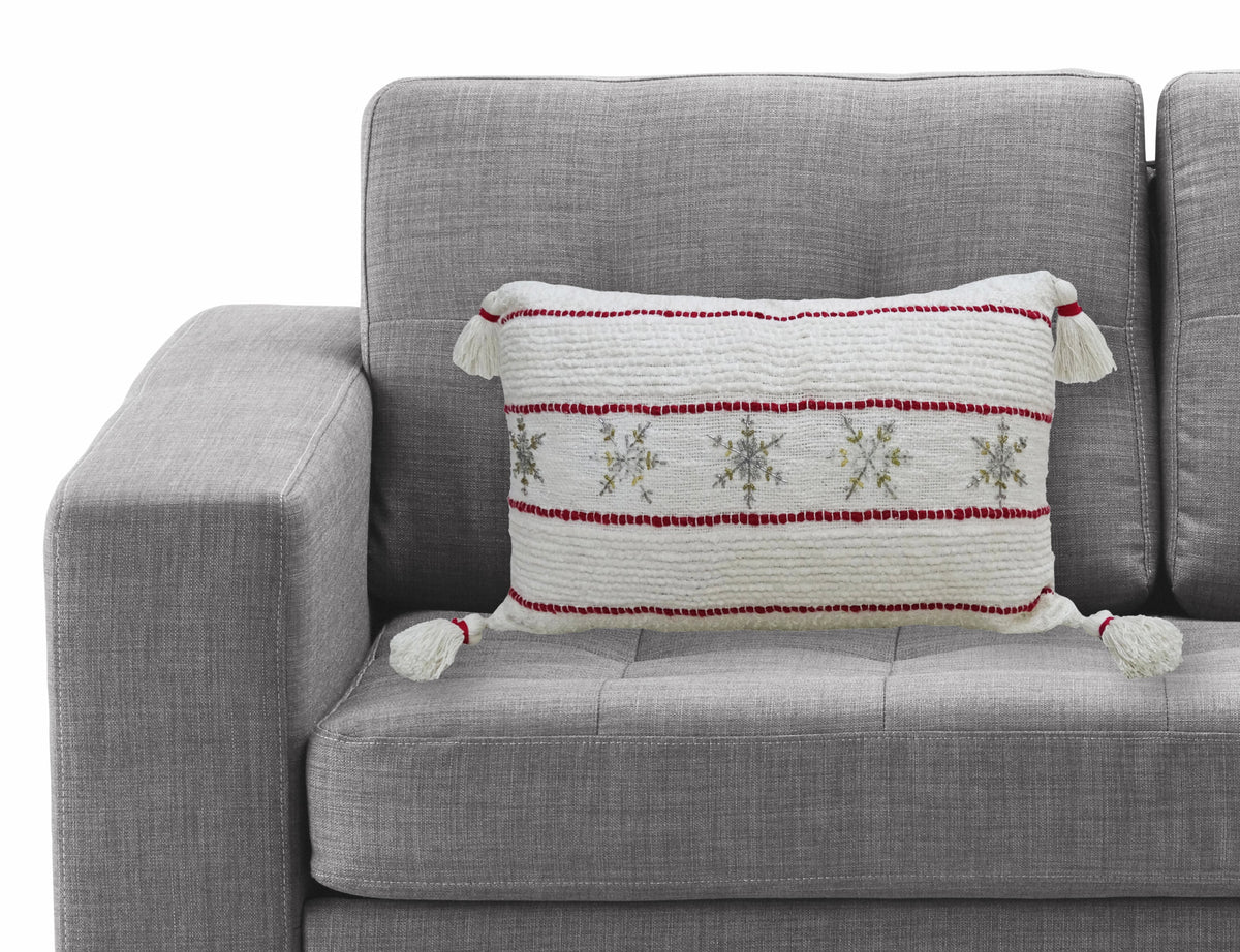 Rectangular white decorative pillow with horizontal red stripes and embroidered silver snowflakes, accented with white tassels on a light gray fabric couch.