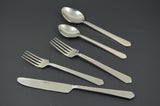 Set of five stainless steel flatware pieces including a serrated knife, two forks, and two spoons with straight, simple handles on a dark surface.