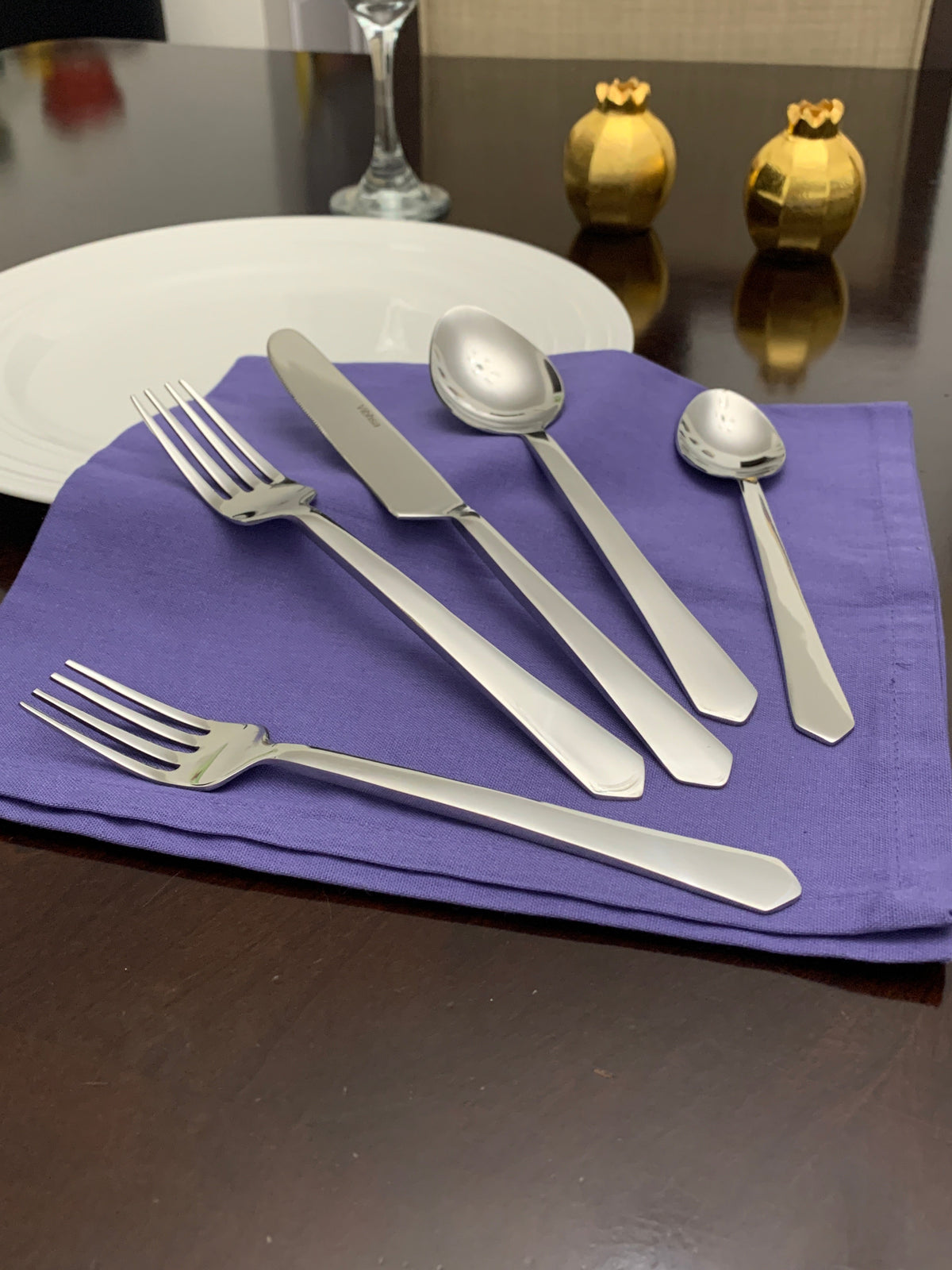 Set of five silver stainless steel flatware pieces including forks, knives, and spoons arranged on a folded purple cloth with a white plate and decorative golden pomegranates in the background.
