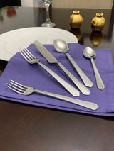 Five-piece silver stainless steel flatware set including two forks, a knife, a tablespoon, and a teaspoon arranged on a purple cloth napkin on a dark wood table.