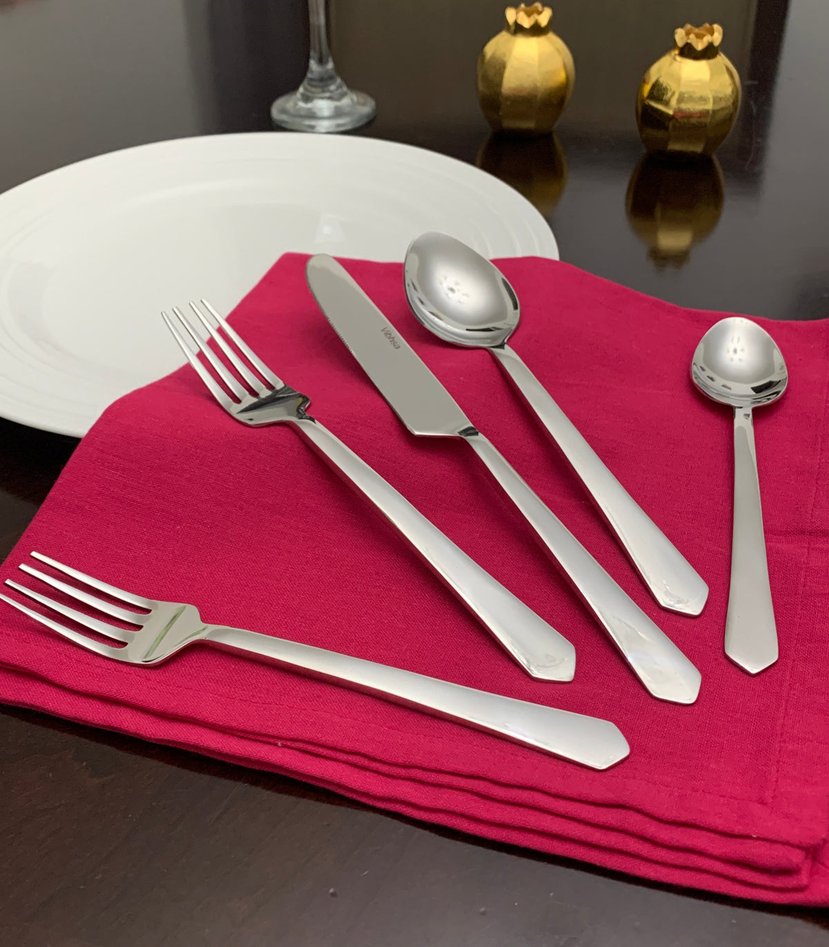 Silver stainless steel flatware set including two forks, one knife, one spoon, and one smaller spoon on a red cloth with a white plate and two gold decorative items in the background.