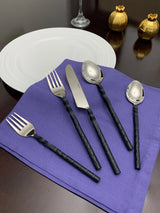 Set of five stainless steel flatware pieces with twisted black handles, including two forks, one knife, and two spoons, displayed on a purple napkin beside a white plate and decorative gold items.