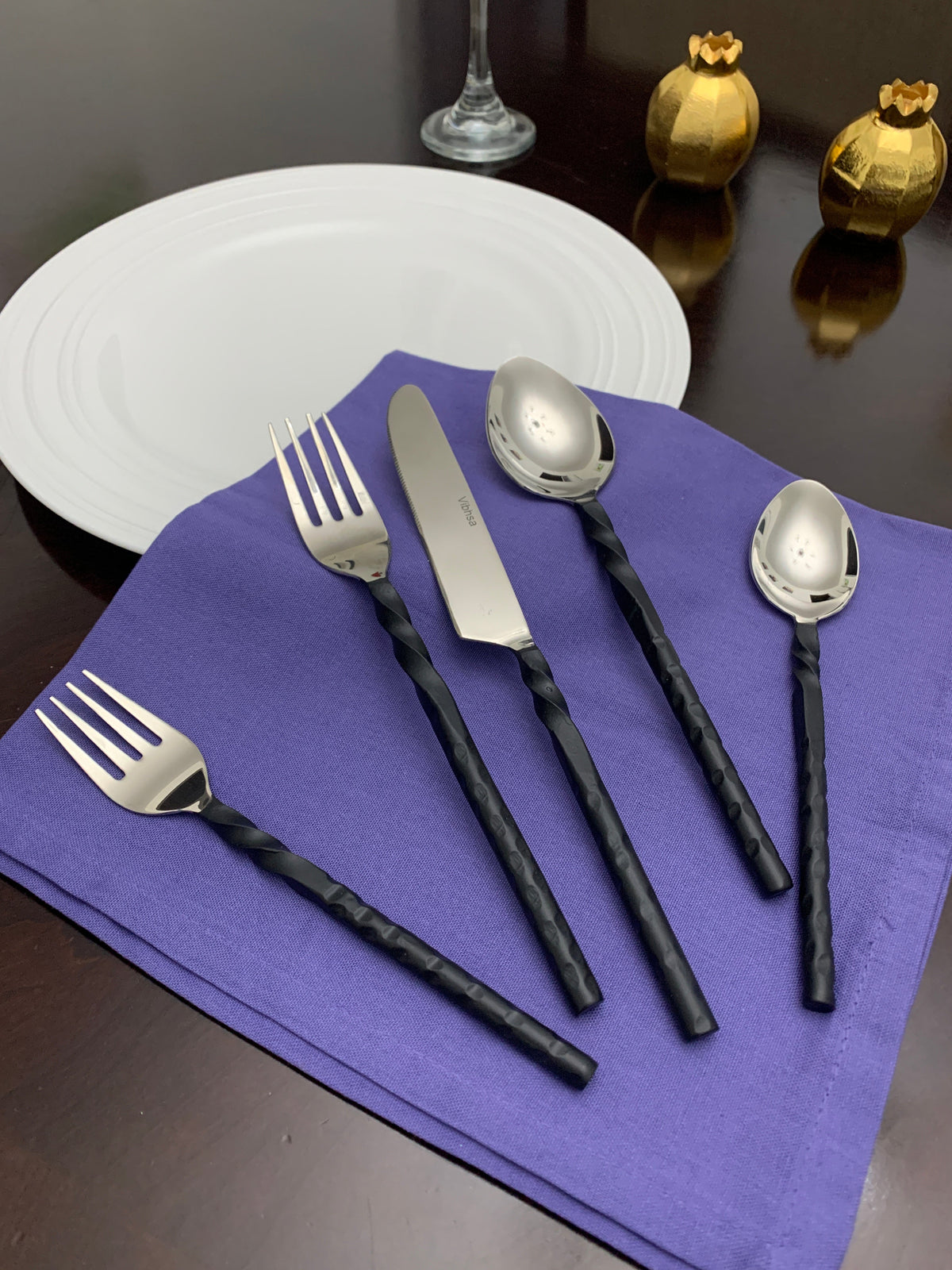 Set of five stainless steel flatware pieces with twisted black handles, including two forks, one knife, and two spoons, displayed on a purple napkin beside a white plate and decorative gold items.