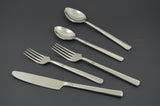 Five-piece stainless steel flatware set including a dinner knife, two forks, and two spoons with simple double-line detail on the handles, shown on a dark background.