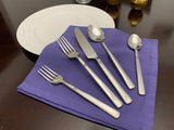 Five pieces of polished silver stainless steel flatware, including two forks, a serrated knife, a tablespoon, and a teaspoon, arranged on a folded purple napkin beside a white plate on a dark wooden table.