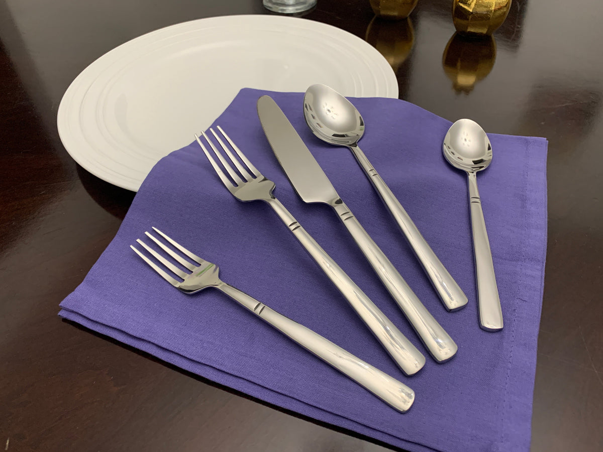 Five pieces of polished silver stainless steel flatware, including two forks, a serrated knife, a tablespoon, and a teaspoon, arranged on a folded purple napkin beside a white plate on a dark wooden table.