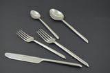 Silver stainless steel flatware including two forks, a knife, a tablespoon, and a teaspoon arranged on a dark gray surface.