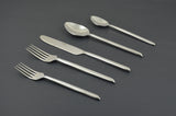 Five-piece stainless steel flatware set including two spoons, a knife, and two forks with sleek, slightly tapered handles on a dark gray background.