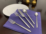 Five-piece stainless steel flatware set with unique sleek handles arranged on a folded purple napkin beside a white plate on a dark wooden table.