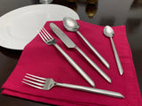Five-piece stainless steel flatware set with uniquely tapered handles arranged on a folded red cloth beside a white plate on a dark table.