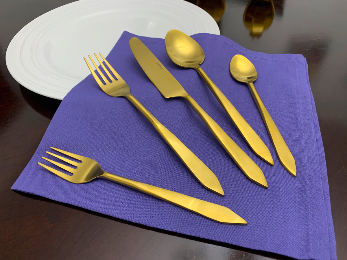 Golden stainless steel flatware set including two forks, a knife, a tablespoon, and a teaspoon, arranged on a purple cloth napkin with a white plate in the background.