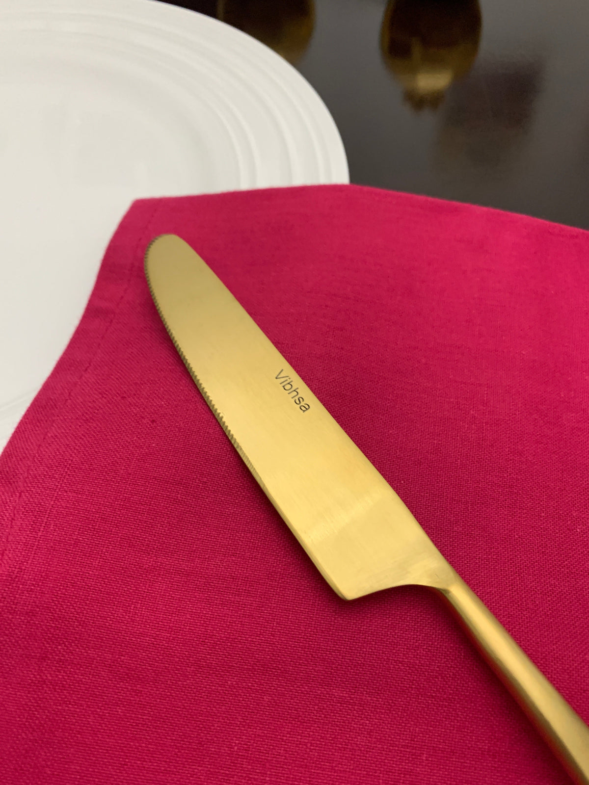 Golden stainless steel butter knife with 'Viksa' branding on the blade, resting on a folded deep pink napkin beside a white plate on a dark reflective surface.