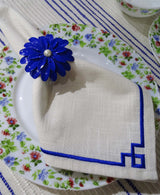 White cloth napkin with blue geometric embroidery and a blue flower-shaped napkin ring with pearl center, placed on a floral-patterned plate with red, green, and blue flowers.